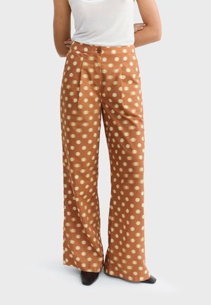 Femme portant un pantalon marron taille haute à jambes larges avec de grands pois beiges, associé à un haut blanc sans manches et des chaussures noires à bouts pointus.