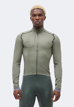Homme aux cheveux blonds courts portant une veste de cyclisme ajustée olive à manches longues avec fermeture éclair et un cuissard rembourré vert foncé.