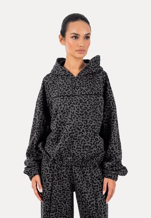 Donna che indossa una felpa con cappuccio grigio scuro a stampa leopardata e pantaloni abbinati, in piedi con le mani rilassate ai lati, guardando leggermente verso destra.