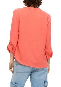 Korallenbluse mit strukturiertem Finish, Dreiviertelärmel mit umgeschlagenen Manschetten und abgerundetem Saum, getragen mit hellblauen Cargo-Hosen.