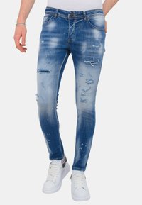 Versleten blauw denim met distressed details, voorzien van scheuren en een licht getailleerde pasvorm, gecombineerd met witte sneakers.