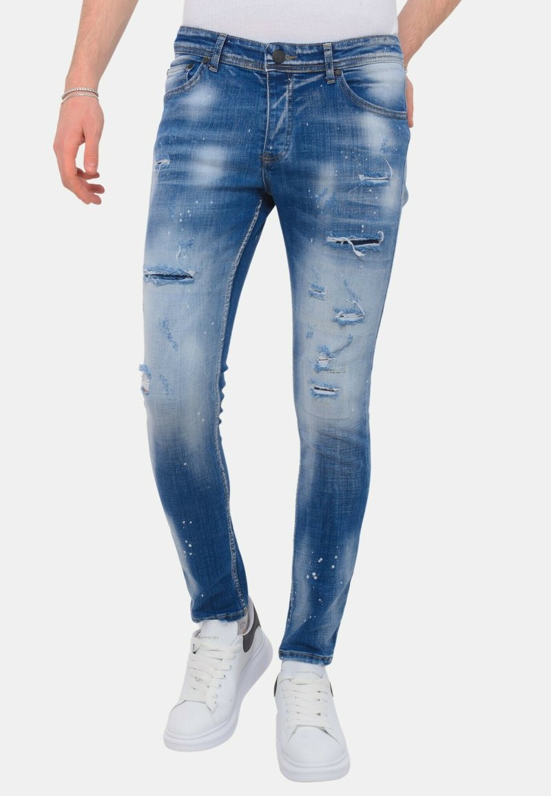 Versleten blauw denim met distressed details, voorzien van scheuren en een licht getailleerde pasvorm, gecombineerd met witte sneakers.