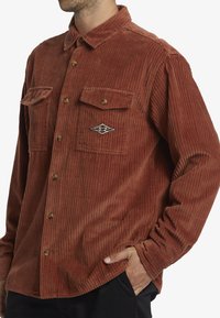 Brown corduroy shirt with long sleeves, two front pockets, rounded collar, button closure, and a diamond-shaped logo on the left chest.