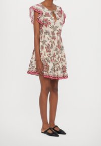 Robe courte florale avec des manches volantées, encolure en trou de serrure et taille froncée. Présente un fond crème léger avec des bordures et des accents rouges.