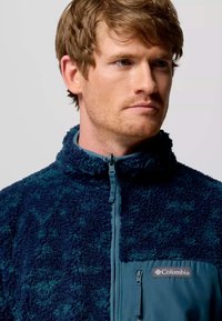 Giacca in pile di colore blu scuro con superficie texturizzata, chiusura frontale con mezzo zip e tasca solida a contrasto. Logo Columbia sulla tasca.