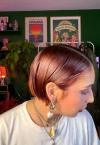 Person with short pink hair wearing silver earrings and layered necklaces, looking down, with green wall and colorful posters in background.