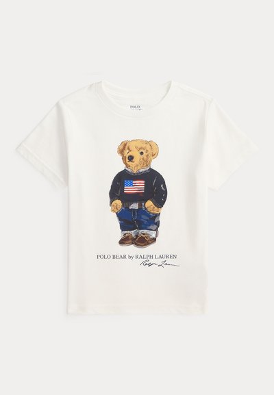 Biały T-shirt z krótkim rękawem z niedźwiedziem ubranym w granatowy sweter z amerykańską flagą, niebieskie dżinsy i brązowe buty, napis Polo Bear by Ralph Lauren.