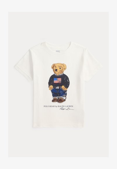 Biały T-shirt z krótkim rękawem z niedźwiedziem ubranym w granatowy sweter z amerykańską flagą, niebieskie dżinsy i brązowe buty, napis Polo Bear by Ralph Lauren.