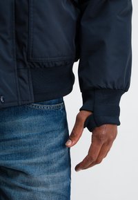 Marineblauer Bomberjacke mit gerippten Bündchen und Saum, ausgestattet mit einem Reißverschluss. Kombiniert mit blauen Jeans aus Denim, die einen strukturierten Materialkontrast zeigen.