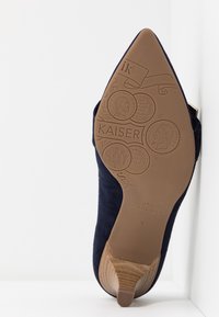 Chaussure à talon haut en suède bleu foncé avec un bout pointu et une semelle en caoutchouc beige ornée de gravures de pièces et de détails de marque.