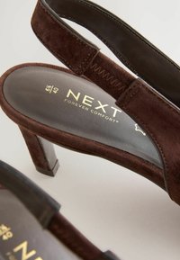Rudos zomšos slingback aukšti kulniukai su tekstūruota dirželiu, lygaus odos akcentu ir kontūruota pado forma. Auksinė logo ant vidpadžio.