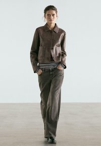 Brun læder kort jakke med to forlommer, knaplukning og spids krave. Kombineret med højtaljede, mørke denimjeans.