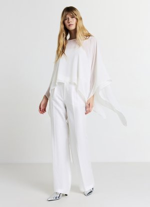 Top en chiffon blanc avec un ourlet asymétrique, assorti à un pantalon en lin blanc fluide. Les chaussures sont pointues en argent métallique. Textures lisses et légères.
