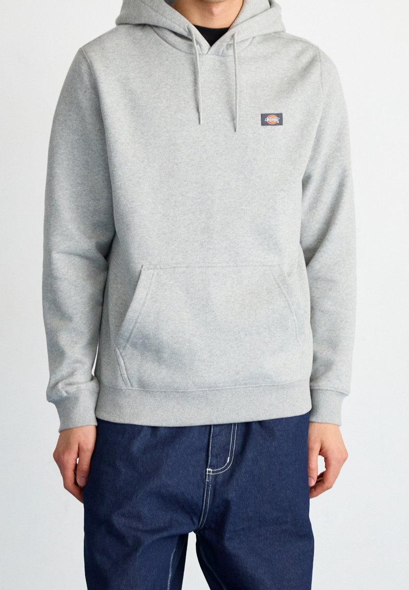 Homme portant un sweat à capuche gris clair avec poche avant et un pantalon bleu foncé sur fond blanc uni.