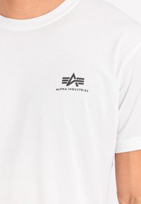 Balta kokvilnas t-krekls ar melnu logotipu, kas attēlo trīs stilizētas spārnus un uzrakstu "ALPHA INDUSTRIES" kreisajā krūšu daļā.