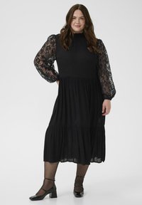 Moteris vilkinti juodą midi suknelę su permatomomis gėlėmis aptrauktomis ilgomis rankovėmis ir juodais blizgiais Mary Jane bateliais, stovi priešais neutralų foną.