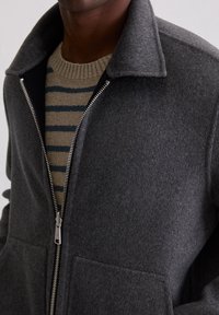 Szara kurtka z mieszanki wełny z zamkiem z przodu, dużymi kieszeniami i kołnierzem koszulowym, nałożona na beżowo-niebieski sweter w paski.