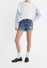 Sudadera blanca con mangas a rayas azules, combinada con shorts de mezclilla deshilachados y mocasines negros con calcetines blancos. Atuendo casual en colores neutros.