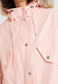 Różowa kurtka z futrzanym kapturem, z metalowymi zatrzaskami i sznurkiem, oraz przednią kieszenią o kanciastym designie.