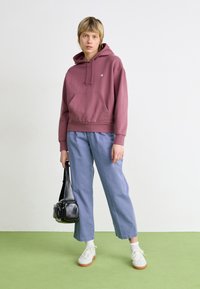 Hettegenser i mauve med kengurulomme, kombinert med lyseblå bukser, hvite sneakers og en svart crossover-veske. Grønn gulv bakgrunn.