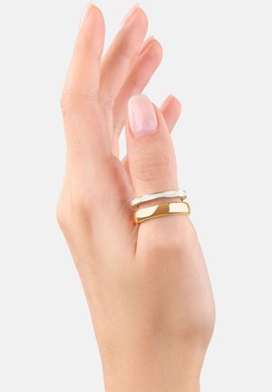 Due anelli su una mano; una fascia d'oro e una fascia d'argento, entrambe lisce con larghezze e finiture leggermente diverse. La pelle è di tonalità chiara.