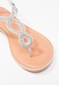 Hopeapärli thong-sandaalit, joissa on äärettömyysdesign, sileä beige pohja ja valkoinen ompelu, korostaen teksturoitua pintaa.