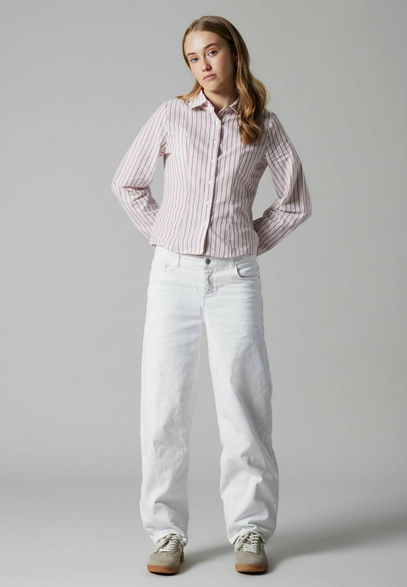 Jeune femme debout, les mains derrière le dos, portant une chemise à rayures roses boutonnée, un pantalon blanc à jambes larges et des baskets beiges.