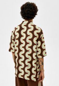 Korte mouwen shirt in bruine stof met een kleurrijk golvend patroon en crème, blauwe en oranje accenten. Label zichtbaar aan de zijkant.