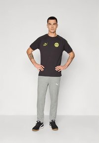 Camiseta negra con detalles y logo en amarillo, combinada con pantalones deportivos grises y zapatillas negras con suela de goma. El modelo está de pie con los brazos a los lados.