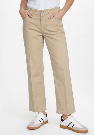 Person trägt beige gerade geschnittene Hose mit Händen in den Taschen und weiße Sneakers mit schwarzen Streifen und Gummisohlen.