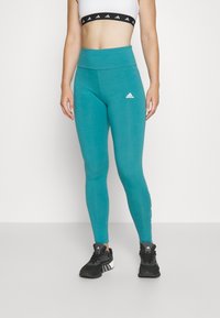 Teal leggings med hög midja, slät textur och en liten vit logotyp på låret, i kombination med svarta träningsskor.