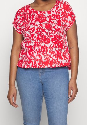 Kvinde iført en rød og hvid blomstrede kortærmet bluse kombineret med lyseblå jeans, stående op ad en ensfarvet baggrund.