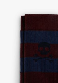 Calcetines a rayas en rojo oscuro y azul con un patrón de calavera y huesos cruzados. Están hechos de un tejido suave y texturizado con puños acanalados.