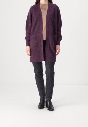 Kobieta ubrana w długi fioletowy kardigan z przednimi kieszeniami, na beżowej bluzce, czarne dżinsy i czarne botki na białym tle.