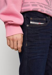 Vaqueros de mezclilla azul oscuro con una etiqueta de bolsillo visible, combinados con un sudadera rosa claro con un diseño gráfico. Textura suave y ajuste casual.