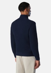 Maglione a costine blu navy con colletto alto, design aderente e motivo a maglia texturizzata, indossato con pantaloni chiari.