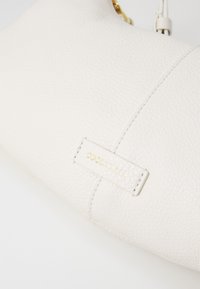 Sac à main en cuir texturé blanc avec une forme incurvée. Présente une plaque logo dorée et des détails de couture minimalistes.
