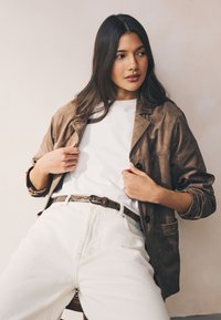 Brown leather jacket with a tailored fit, white t-shirt, and high-waisted white jeans, accented by a patterned belt and relaxed pose.