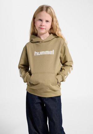 Młoda dziewczyna z długimi blond włosami ubrana w khaki kaptur marki Hummel i ciemne dżinsy, stoi z rękami w kieszeniach bluzy na tle białego tła.