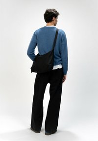 Schwarze Nylon-Schultertasche mit einer geschwungenen Form, verstellbarem Tragegurt und glatter Textur. Getragen von einer Person in einem blauen Pullover und schwarzen Hosen.