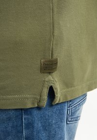 Oliwkowa bawełniana koszulka z teksturowaną powierzchnią, bocznymi rozcięciami i małą brązową metką z logo na dolnym zakończeniu. Zestawiona z niebieskimi dżinsami.