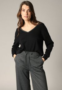 Pull noir à col en V avec des manches longues, porté avec un pantalon gris à rayures avec taille à cordon de serrage et poches avant.