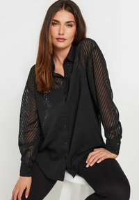 Blusa nera trasparente con motivo a spina di pesce, chiusura con bottoni sul davanti e maniche lunghe. Il tessuto strutturato aggiunge profondità, abbinata a pantaloni neri aderenti.