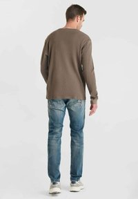 Hombre de pie, de espaldas a la cámara, vistiendo una camiseta de manga larga marrón, jeans azules y zapatillas gris y blancas.