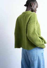 Femme aux cheveux tressés portant des boucles d'oreilles argentées en forme de créoles, un pull vert duveteux et un jean bleu clair, de profil contre un fond blanc uni.