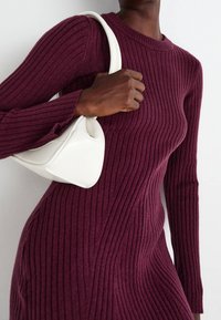 Žebrované šaty s dlouhým rukávem v tmavě burgundské barvě, doplněné malou bílou kabelkou s zakřiveným designem, kterou drží štíhlá ruka.