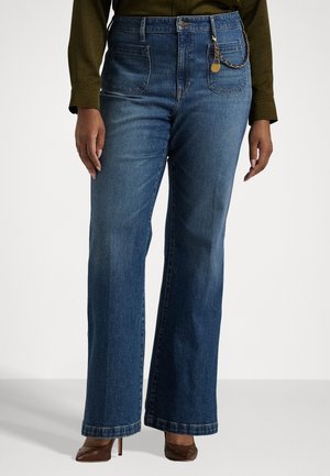 Vrouw met een high-waisted, wijd uitlopende blauwe spijkerbroek met voorzakken en gouden kettinkdetail, bruine hakken en een olijfgroen longsleeve top.