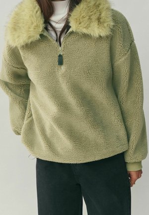 Kobieta w jasnozielonym pulowerze sherpa z meszkowanym kołnierzem, półzamkiem z przodu, ściągaczami na rękawach i czarnych spodniach na jednolitym tle.