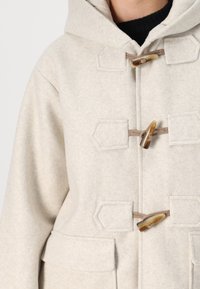 Beige ullfrakk med hette, hempefester og to frontlommer. Stoffet har en myk, teksturert overflate.