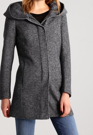 Femme portant un manteau gris ajusté et texturé avec un large col sur un jean bleu, debout devant un fond clair uni.